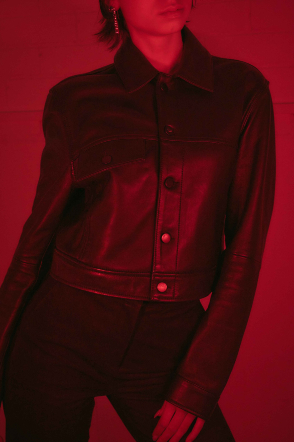 In red lighting, a women model wears the Brut Female Ape Cropped black leather jacket